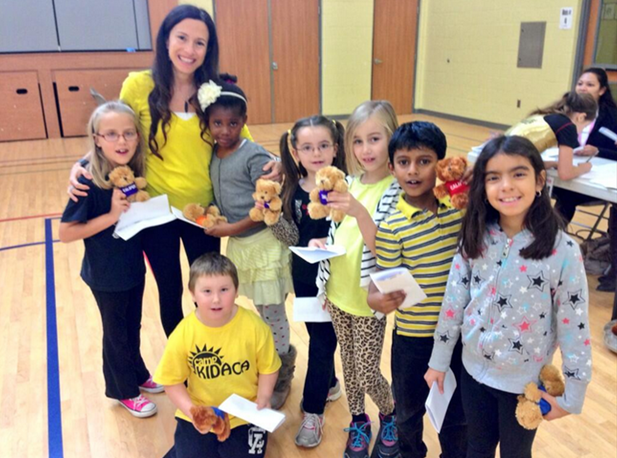 A group of school-age children stand smiling in a group with their teacher in a school gymnasium, all holding teddy bears